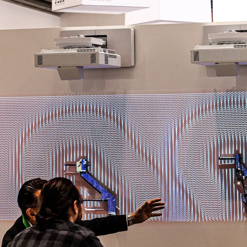 A trade fair visitor gets informed by an employee of the trade fair stand about the product on offer, which is illuminated by a projection