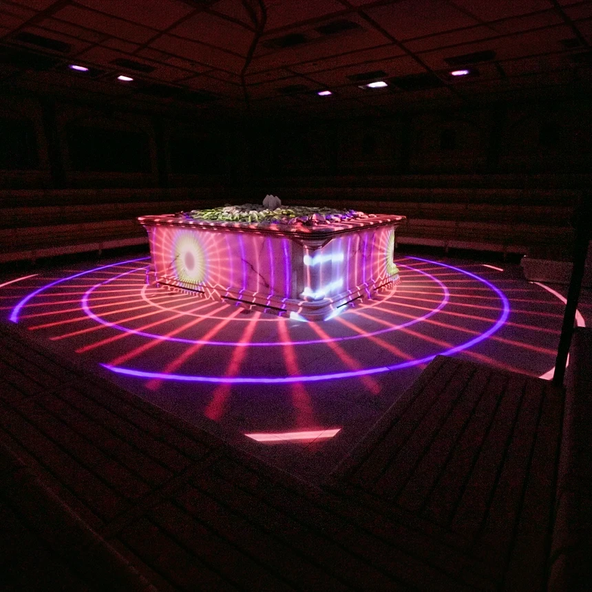 Energetic light projection in the Crystal Palace: Concentric rings and radial beams in pink and violet circle the sauna heater—large-scale projections, light, and music create an immersive *Aufguss* experience by TNL GmbH.