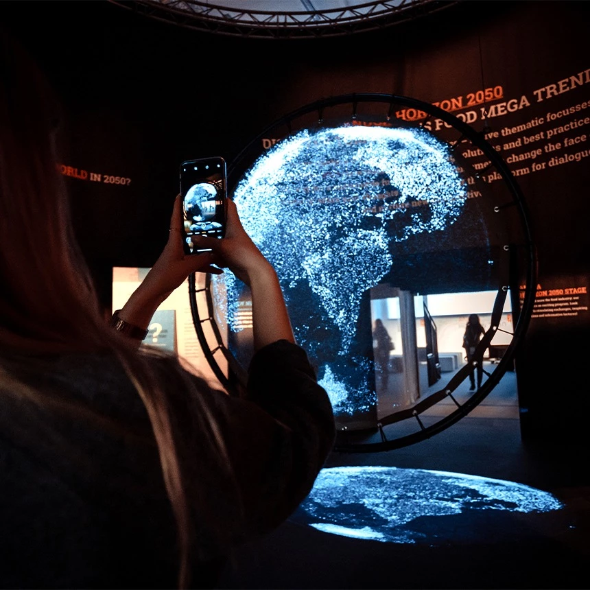 Circular projection with augmented reality effect: a globe is shown as a holographic 3D installation in the showroom at the Cologne trade fair, with a visitor in front of it taking a picture with her cell phone