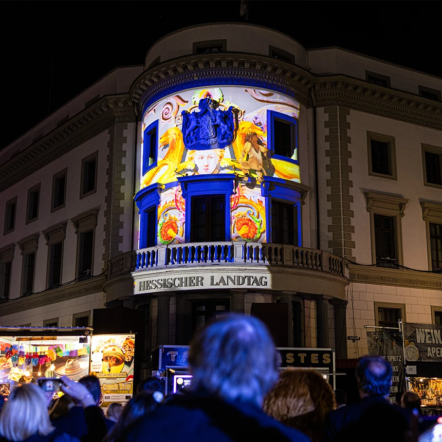 Die Fassade des hessischen Landtags wird mit einer bunten Illumination und blau umrahmten Fenstern gezeigt. Der Löwe steht mit dem Rücken zum Publikum.