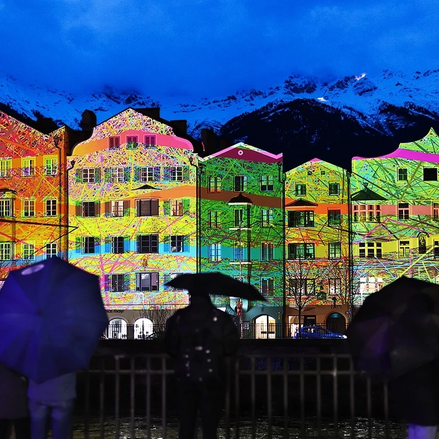 Three people stand in front of the Maria Hilf row of houses in Innsbruck, which is illuminated with a colorful video projection for each building