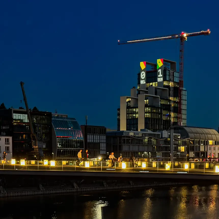 Fassadenprojektion an den UNIQ TOWERS im Düsseldorfer Medienhafen zeigt den Live-Spielstand Belgien gegen Senegal, im Vordergrund Passanten auf der Kaipromenade.