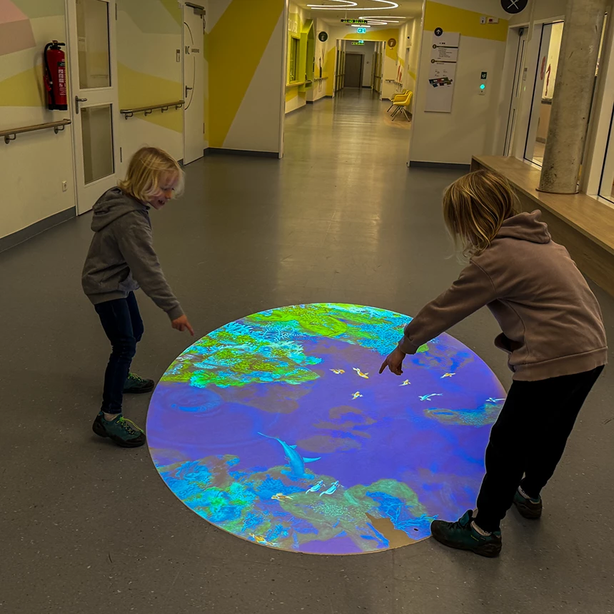 2 Kinder zeigen auf eine interaktive Projektion auf dem Boden in der Kinderklinik Bethel, auf der bunte Fische und Korallen zu sehen sind