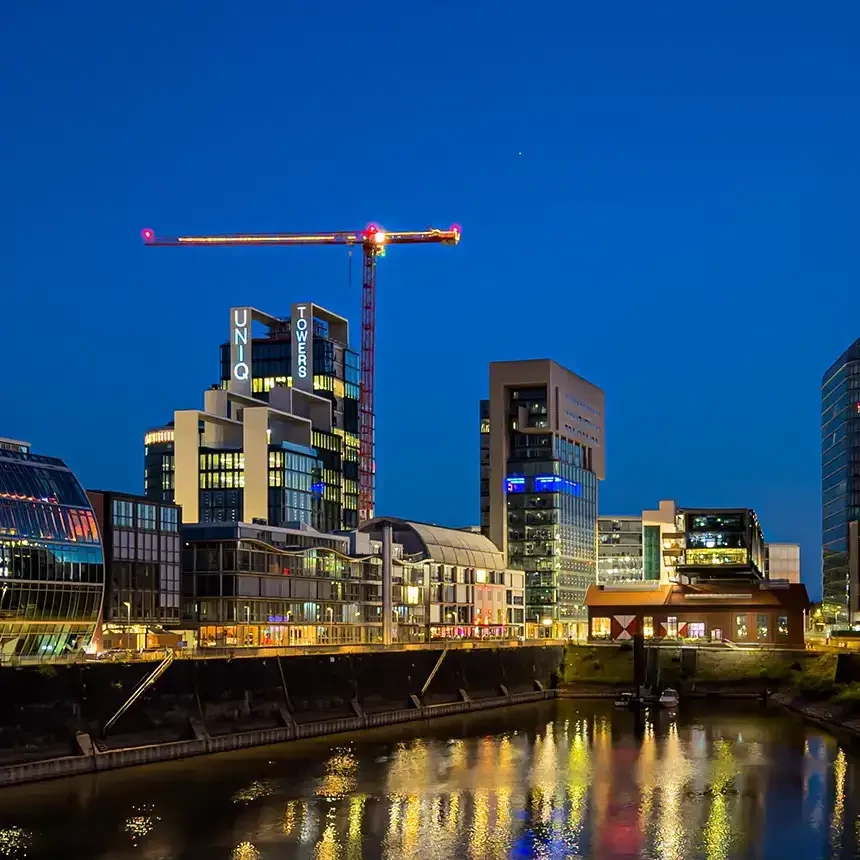 Blick auf die UNIQ TOWERS am Düsseldorfer Medienhafen: Logoprojektion auf der Fassade der Türme des Gebäudes, außderdem ist ein Baukran und Lichtreflexionen im Hafenbecken zu sehen.