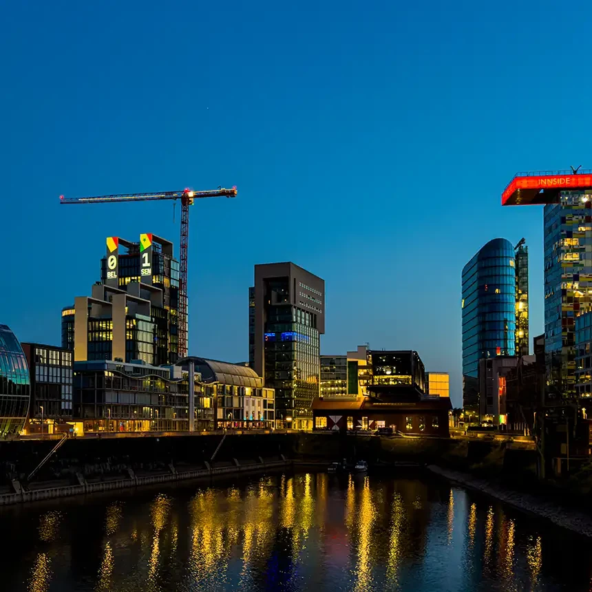 Weitwinkelaufnahme des Medienhafens Düsseldorf: Zwischen Kranstruktur und angrenzender Bebauung zeigt die Fassadenprojektion der UNIQ TOWERS den aktuellen WM-Spielstand, während sich die Lichter im Wasser spiegeln.