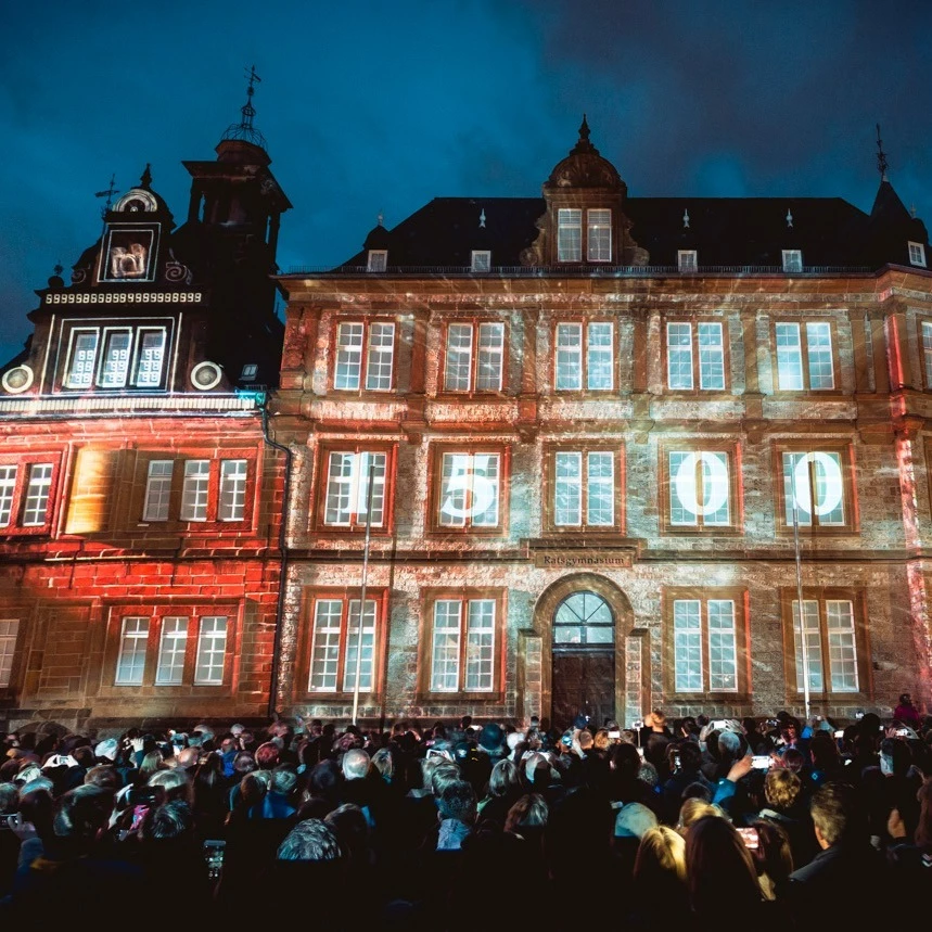 A crowd of people watches a projection mapping projected in red onto a facade