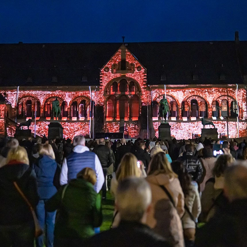 Zahlreiche Besucher:innen schauen die Projektionsshow in Goslar und dokumentieren sie mit Ihren Handys, auf dem Gebäude sind Pflanzen und eine dörfische Szene dargestellt