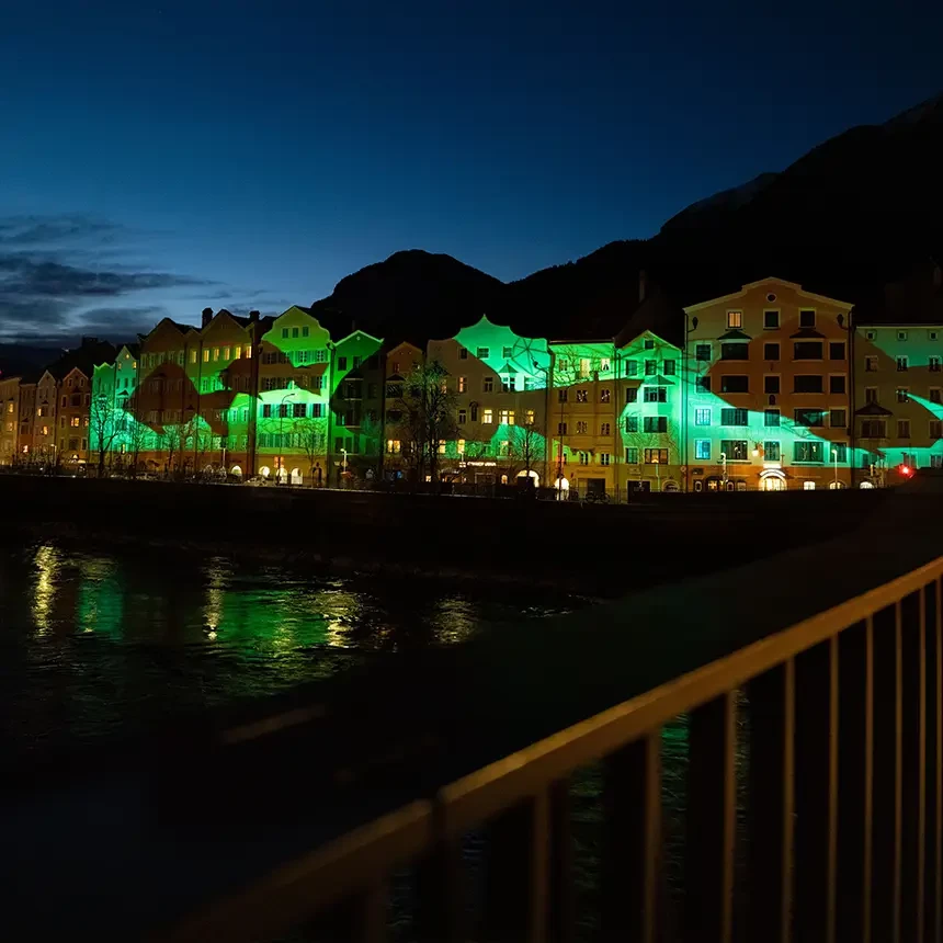 Illumination der Innsbrucker Mariahilf-Häuserzeile mit dem Inn im Vordergrund und Alpenpanorama im Hintergrund: es sind Silhouetten von Fischen im Origami-Stil zu sehen