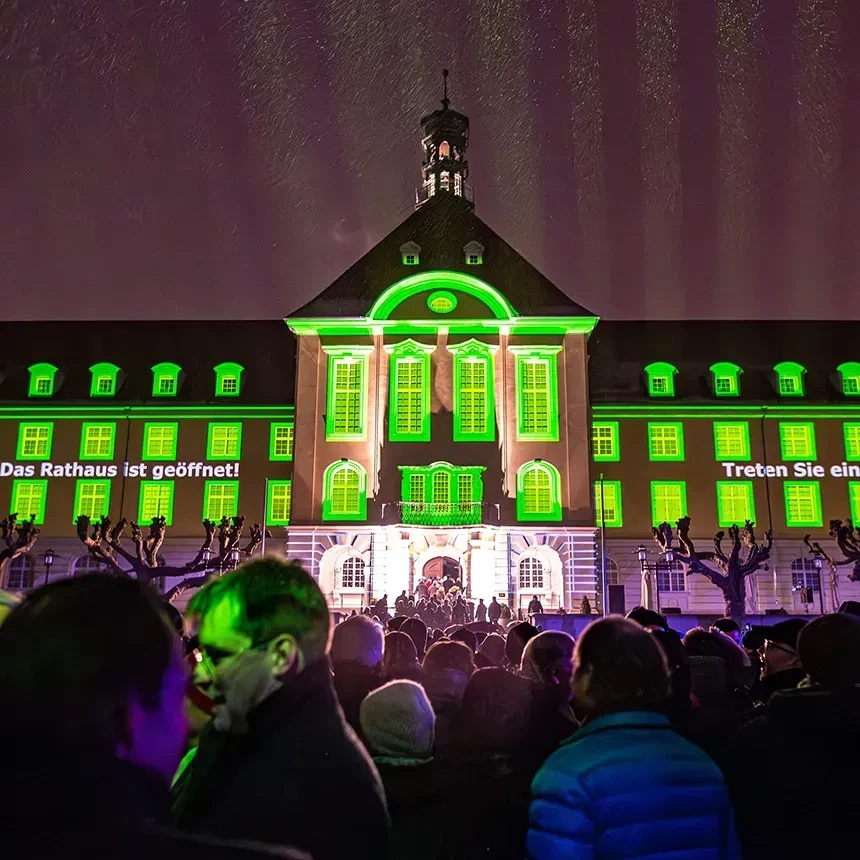 A projection illuminates the windows of a town hall in green colors and the entrance is illuminated with bright white light
