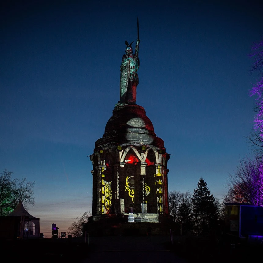 Object mapping with red and yellow industrial effects on the Hermann monument: gears and chain hoists are projected onto the monument in the area of the columns