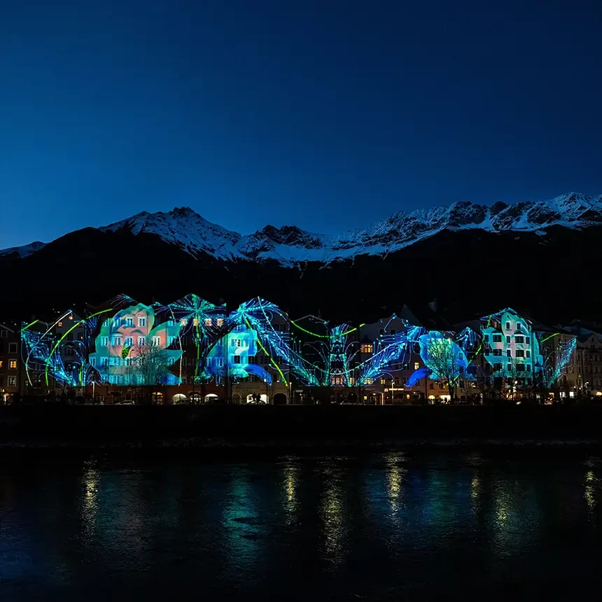3D Video Mapping mit floralen Motiven auf die Skyline der Innsbrucker Mariahilf-Häuserzeile mit dem Inn im Vordergrund und Alpenpanorama im Hintergrund